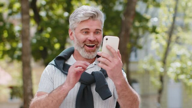 Happy Caucasian bearded man use mobile smartphone celebrating win good message news, lottery jackpot victory, online outdoors. Male middle-aged freelancer in city street. Business people lifestyle.
