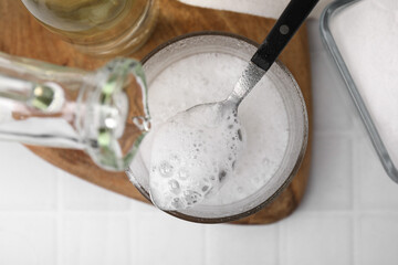 Pouring vinegar into spoon with baking soda over bowl at white tiled table, top view