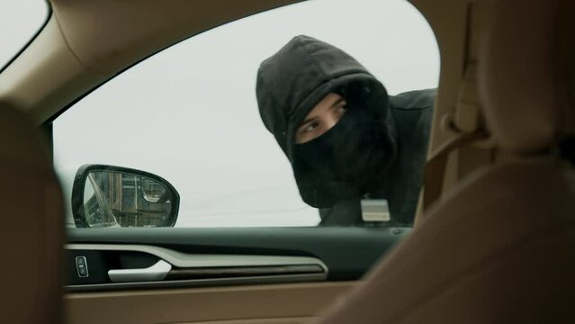 Hooded thief in black breaks into car criminal act vehicle theft auto crime in progress. Person in black hood hacks car lock car crime auto theft vehicle criminal activity man thief.