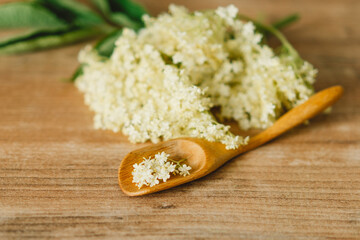 Freshly picked elderflower blossoms lie on a rustic wooden table, with a small wooden spoon holding a few delicate flowers, bathed in natural light.