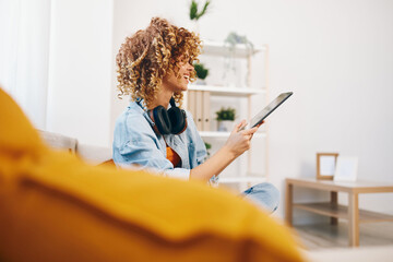 Home-reading Woman: A Happy, Smiling Gamer Girl Sitting on a Cozy Couch, Holding a Tablet PC, Enjoying Online Games and Chatting with a Friend via Headphones, Surrounded by a Beautifully Decorated