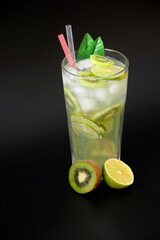 A tall faceted glass of fruit juice with ice and straws on a black background, next to pieces of ripe kiwi and lime.