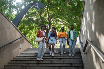 Cheerful, cohesive group of African American students enters subway underpass together, heading to studies as team. Happy, joyful black friends girl and guy down city stairs walking, chatting, smiling