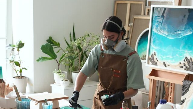 Female artist pours epoxy resin and smears it with stick on wooden board. Craftswoman using liquid art technique, creating picture of sea and waves, closeup view. Art deco, master class on resin art. 