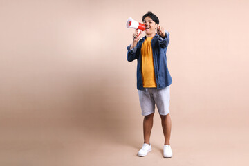 Full Body Of Young Asian Man Shouting On Megaphone While Pointing to You Isolated On Beige Background