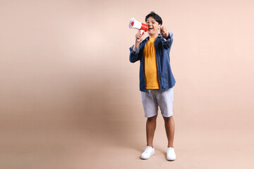 Full Body Of Young Asian Man Shouting On Megaphone While Pointing to You Isolated On Beige...