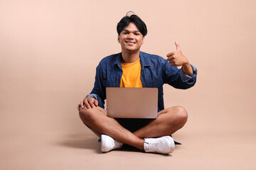 Full Length Of Excited Young Asian Man Giving Thumbs Up While Sitting And Using Laptop Isolated On...