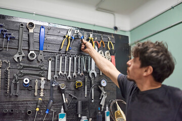 Hispanic repairman grabbing a cable cutter from a tool board in the wall of a bicycle repair shop....