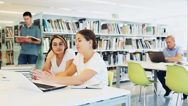 Two positive ladies wearing casual clothes behave loudly while studying in the library and using computer