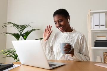 Young Black woman waving hand on a video call at home office using wireless earphones and laptop