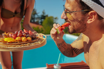 Man eating watermelon by a pool and a woman with a tray of fruits and snacks