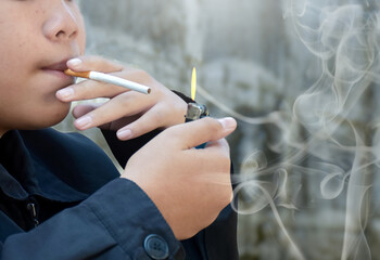 A young male teenager is holding a lighter to light the tip of a cigarette to try smoking it,...