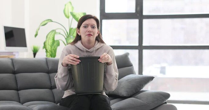 Young woman sits on sofa and vomits into bucket
