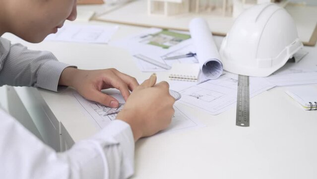 Close up of skilled architect engineer hand draws blueprint on meeting table with blueprint, architectural equipment, house model and safety helmet scatter around. Focus on hand. Immaculate.