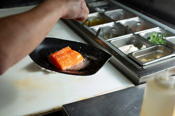 A view of a cook seasoning a raw salmon filet, in a restaurant kitchen setting.