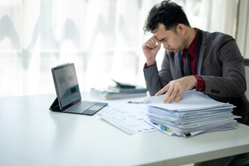 A man is sitting at a desk with a laptop and piles of papers