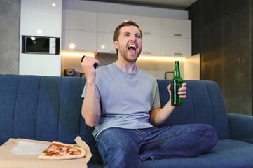 Happy young man drinking beer and eating pizza when watching game on tv at home