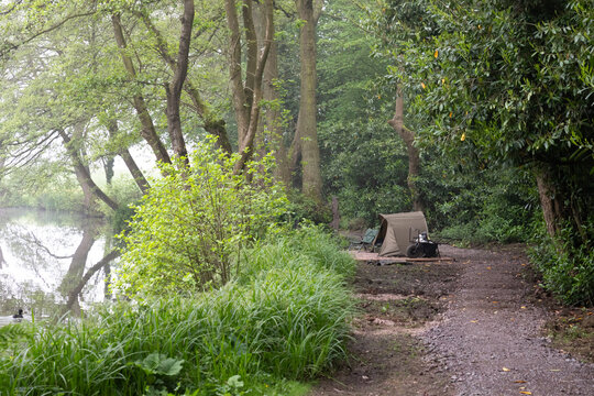 Looking down a footpath by the side of a pool is a fishing set up. A bivvy is pitched by the pool with wheelbarrow and chair. No people and space for text