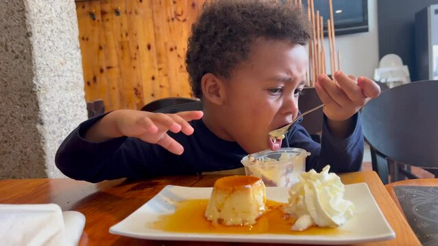 Black kid 4y.o enjoying  an ice cream seated inside a cozy restaurant. He is very expressive and  wears black curly hair. 4K 60FPS