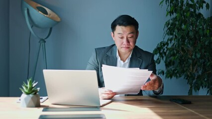 Tired japanese entrepreneur falling asleep while reading boring documents during work process in sunny cabinet. Hardworking male boss reviewing paper reports and preparing business project indoors.