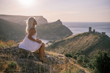 A woman is sitting on a hillside overlooking the ocean. She is wearing a white dress and has blonde hair. The scene is serene and peaceful, with the ocean in the background.