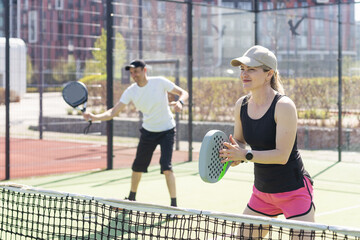 Portrait of positive young woman and adult man standing on padel tennis court, holding racket and ball, smiling 