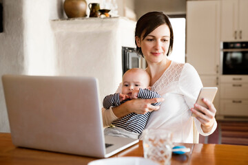 Mother with small baby working from homeoffice, typing on laptop. Mom paying bills online, holding...