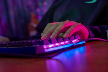 Professional online gamer hand fingers mechanical keyboard in neon color blur background. Soft focus, back view.