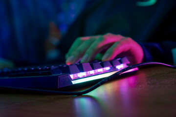 Professional online gamer hand fingers mechanical keyboard in neon color blur background. Soft focus, back view.