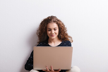Portrait of gorgeous teenage girl sitting, typing on notebook. Studio shot, white background with...