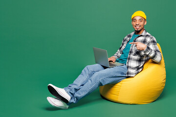 Full body young man of African American ethnicity he wear shirt blue t-shirt yellow hat sit in bag chair hold use work point on laptop pc computer isolated on plain green background Lifestyle concept