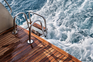 Back teak wooden deck and metal ladder of a of motor yacht, waves and sea splashes and foam.