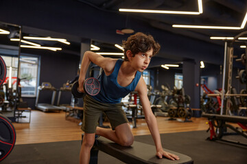 Sportive preteen boy exercising with heavy dumbbells at gym