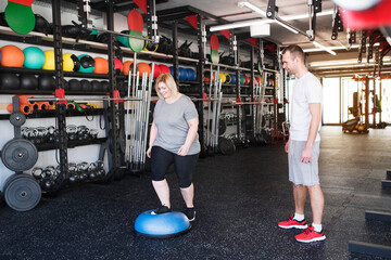 Overweight woman exercising on balance ball in gym. Personal trainer couching her and helping her.