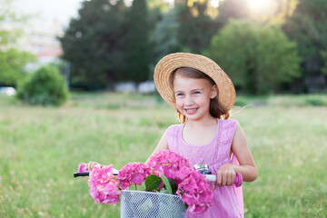 Kid riding bike in park. Child girl in straw hat having fun, smiling at sunset. Pink flowers in...