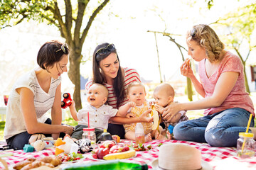 Mothers and babies enjoying group picnic outdoor in park, sitting on picnic blanket and preparing food and drinks.