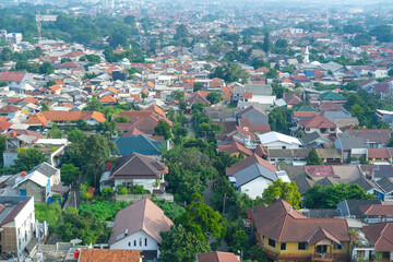 A panoramic view of Bogor city with densely packed houses, green trees, and distant mountains covered in light mist, showcasing the balance of urban life and natural beauty