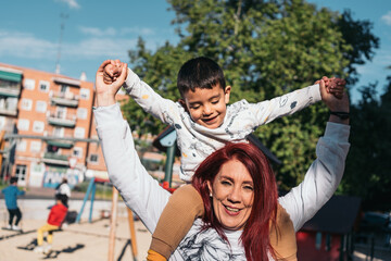 Autistic child riding on mother's shoulders at park. Happy young boy plays with his smiling mother...