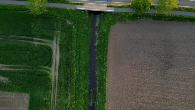 Abstract aerial top-down shot follows a small creek in the soft evening light of Germany. A car cosses the and geometric landscape.