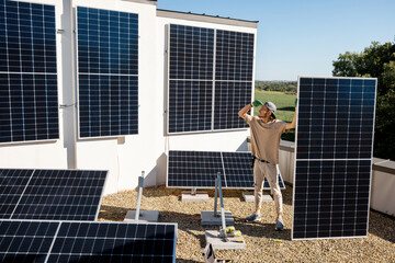 Portrait of a man standing with solar panel on a rooftop of his house during installation process, wide angle view. Owner of property installing solar panels for self consumption