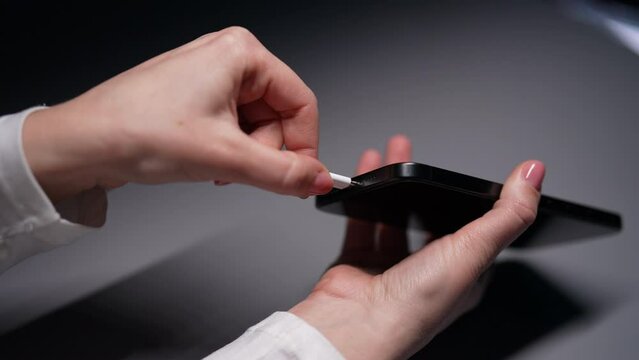 Closeup cropped shot of unrecognizable woman plugging type-c cable into smartphone connector to charge at desk. Closeup female hands connecting cable for transfer data in room with dark interior.
