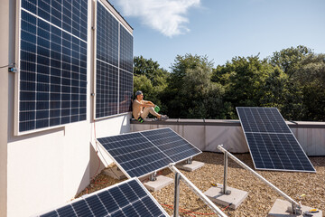 Man sitting on a rooftop of his house with a solar power station installed on it. Renewable green...