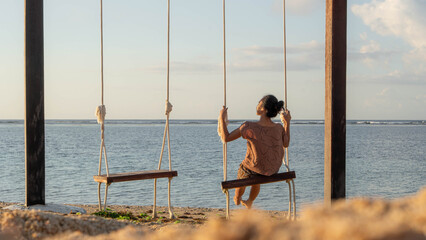 A woman is seen from behind, swinging over the ocean as the sunsets on the beach.