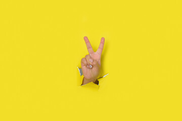 Hand making a sign of victory, peace, coming out of the hole in a torn yellow paper background.