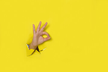 Hand making a sign of OK, coming out of the hole in a torn yellow paper background.
