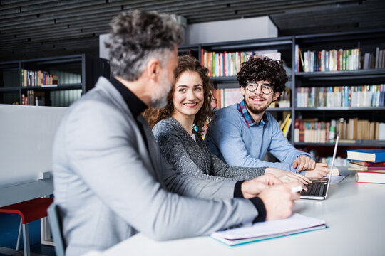 Handsome university professor helping students with final project, showing them research paper, book for thesis in library.