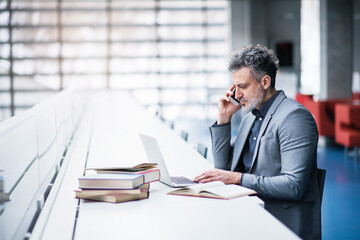 Handsome businessman working on laptop, searching for informations in book and making phone call....