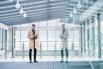 Handsome businessman standing in modern office hallway, holding smartphone in hands.