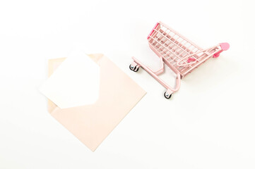 Top view of pink shopping trolley on white background. Pink envelope, white card, post. Creative...