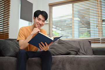 Handsome asian man reading book on couch at home. Hobby, leisure and knowledge concept